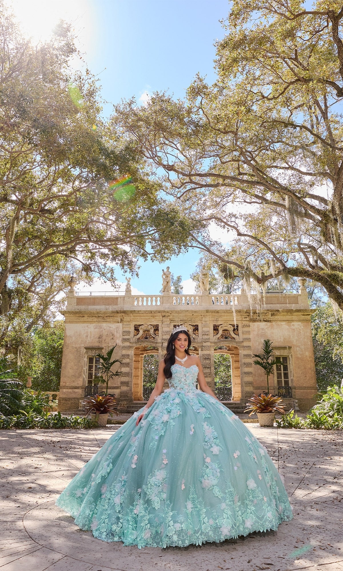 A young woman in the Floral Mint Quinceanera Dress: Princesa PR30133 with delicate floral embroidery stands outdoors before a historic stone building surrounded by large trees.